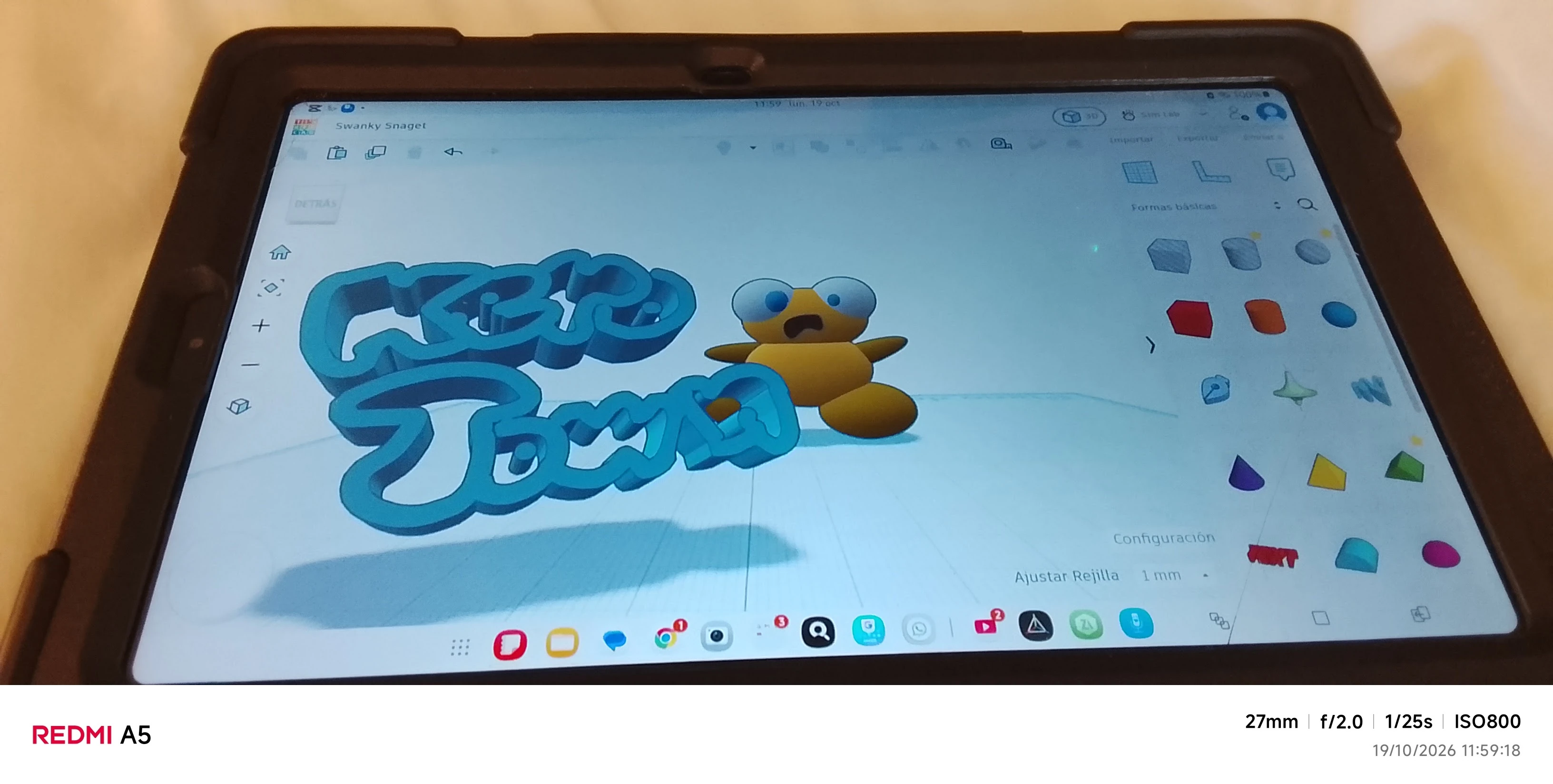 A photo of a tablet in a dark rubber case lying on a cream sheet, its screen showing a 3D design app with a pale blue grid. On the left, thick blue bubble letters in two rows read Kero Town, seen at an angle, with their shadow on the grid. On the right stands a 3D Kero built from smooth golden shapes: a round head with two big white eyes with blue pupils, a dark wavy open mouth, a round body, pointed arms held out to the sides and two big oval feet. A panel of coloured basic shapes runs down the right side of the screen and a row of app icons along the bottom. Below the photo, a white camera strip gives the phone’s model, the camera settings and the date and time.