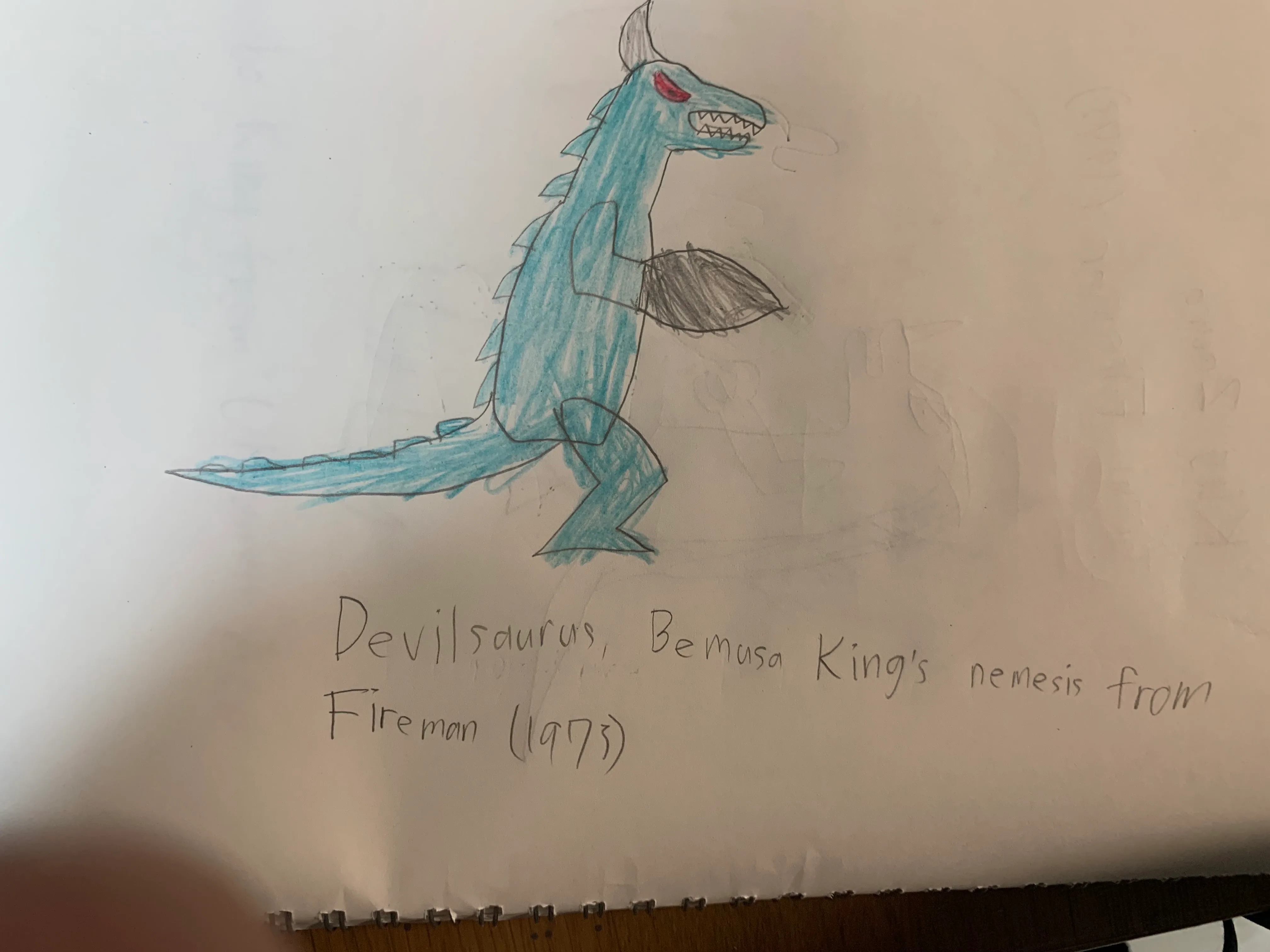 A coloured-pencil drawing on white sketchbook paper of Devilsaurus, a blue dinosaur kaiju standing upright in profile, with a red eye, jagged teeth, a grey horn, spikes down its back and tail and a large grey fin on its arm, above the handwritten caption Devilsaurus, Bemusa King’s nemesis from Fireman (1973).