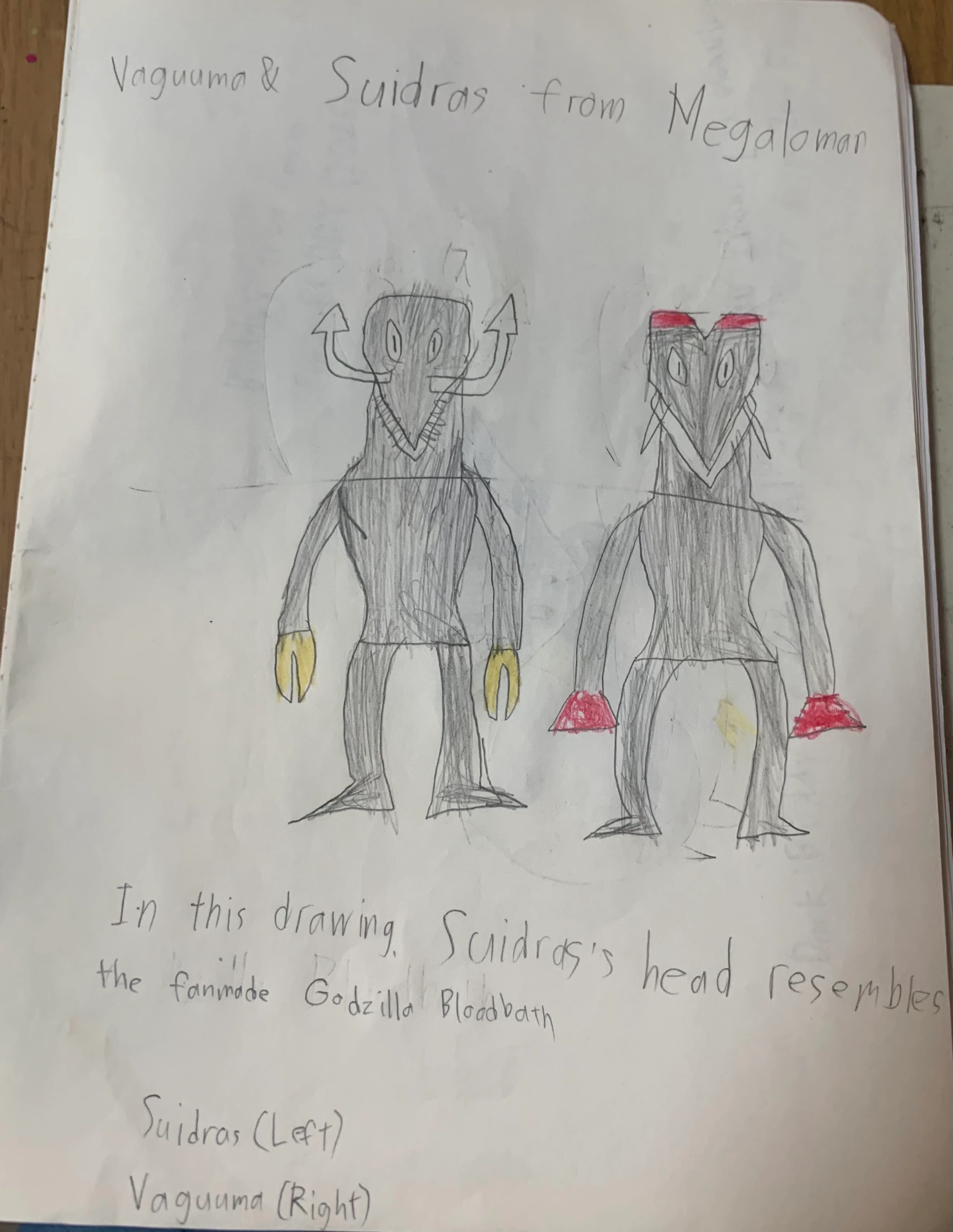 A photo of a pencil drawing on a notebook page lying on a wooden desk. The handwritten heading reads Vaguuma & Suidras from Megaloman. Below it two grey shaded kaiju stand side by side. On the left, Suidras has a tall flat-topped head with two oval eyes, a V-shaped jaw lined with small teeth, two hooked horns ending in arrowheads that curve up from the sides of its head, and yellow pincer claws. On the right, Vaguuma has two red crests on top of its head, a pointed face with oval eyes and thin spikes at its cheeks, and red claws. Underneath, a handwritten note compares Suidras’s head to a fan-made Godzilla, above the labels Suidras (Left) and Vaguuma (Right).