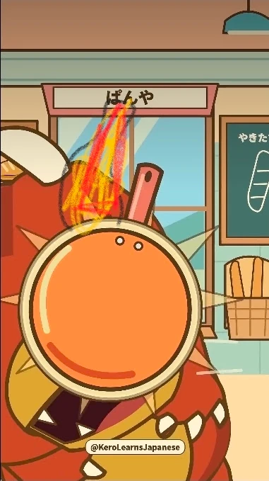A frame from a Kero Learns Japanese Short in a bakery under a ぱんや (bread shop) sign, with a chalkboard and a basket of bread by the window. A big orange frying pan covers the face of Goro, a red spiky monster whose open mouth with white teeth shows below it, and flames have been scribbled above the pan in red, yellow and grey crayon. The channel’s @KeroLearnsJapanese label sits at the bottom.